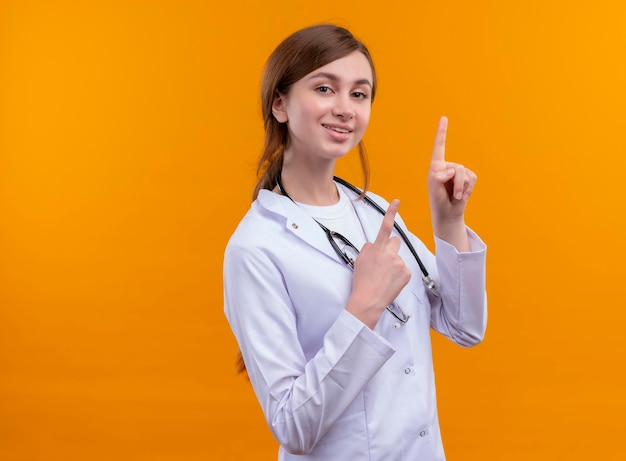 Impresionada joven doctora vistiendo bata médica y estetoscopio apuntando hacia arriba en el espacio naranja aislado con espacio de copia