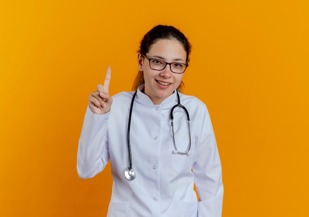 Impresionada joven doctora vistiendo bata médica y estetoscopio con gafas apunta hacia arriba aislado