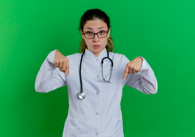 Impresionada joven doctora vistiendo bata médica y estetoscopio y gafas apuntando hacia abajo aislado en la pared verde