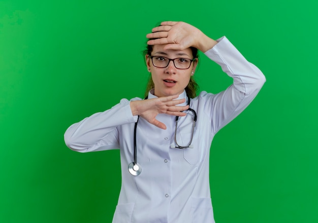 Impresionada joven doctora vistiendo bata médica y estetoscopio y gafas manteniendo las manos debajo y en la cabeza aislada en la pared verde