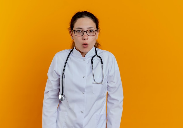 Impresionada joven doctora vistiendo bata médica y estetoscopio y gafas mirando aislado