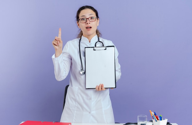 Impresionada joven doctora vistiendo bata médica y estetoscopio y gafas de pie detrás del escritorio con herramientas médicas mirando levantando el dedo mostrando el portapapeles aislado