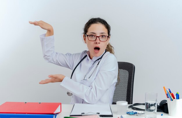 Impresionada joven doctora vistiendo bata médica y estetoscopio y gafas sentado en el escritorio con herramientas médicas mirando haciendo gesto de tamaño aislado