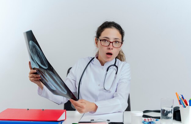 Impresionada joven doctora vistiendo bata médica y estetoscopio y gafas sentado en el escritorio con herramientas médicas sosteniendo una toma de rayos x mirando aislado