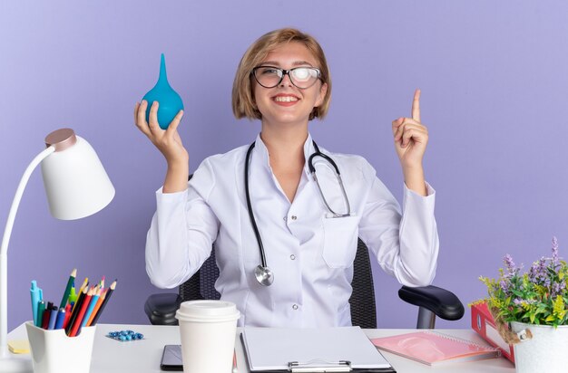 Impresionada joven doctora vistiendo bata médica con estetoscopio y gafas se sienta a la mesa con herramientas médicas sosteniendo enema y apunta hacia arriba aislado en la pared azul