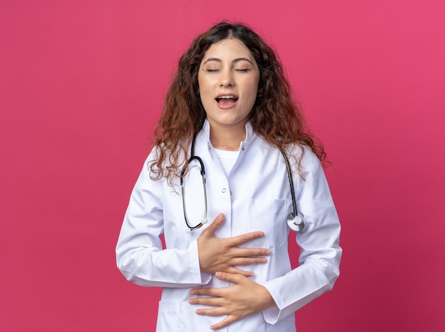 Impresionada joven doctora vistiendo bata médica y estetoscopio manteniendo las manos sobre el vientre con los ojos cerrados aislados en la pared rosa