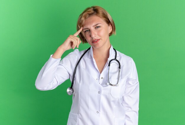 Impresionada joven doctora vistiendo bata médica con estetoscopio poniendo el dedo en la sien aislado en la pared verde