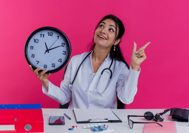 Impresionada joven doctora vistiendo bata médica y estetoscopio sentado en el escritorio con herramientas médicas sosteniendo el reloj girando de cabeza a lado mirando hacia arriba y levantando el dedo aislado en la pared rosa