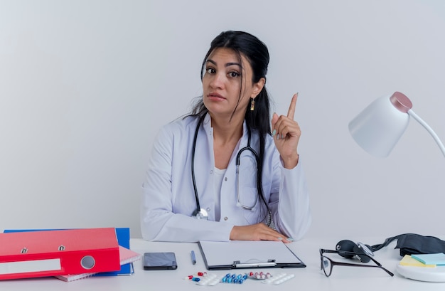 Impresionada joven doctora vistiendo bata médica y un estetoscopio sentados en el escritorio con herramientas médicas mirando poniendo la mano sobre el escritorio levantando el dedo aislado