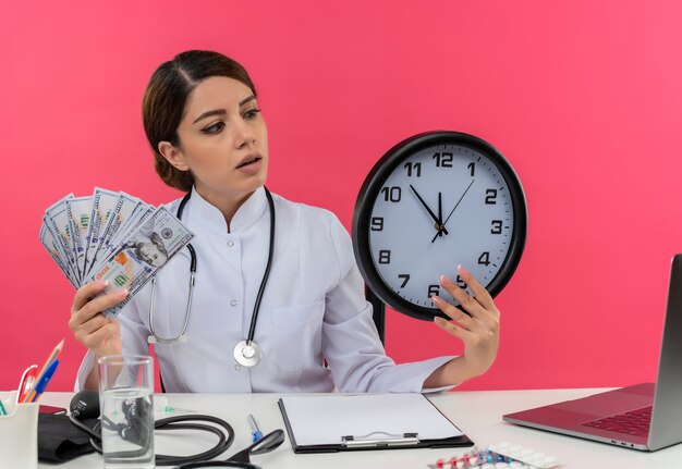 Impresionada joven doctora vistiendo bata médica y un estetoscopio sentados en el escritorio con herramientas médicas y portátil con dinero y reloj mirando el reloj