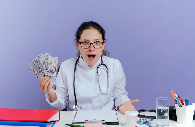 Impresionada joven doctora vistiendo bata médica y un estetoscopio sentados en un escritorio con herramientas médicas sosteniendo dinero mirando aislado