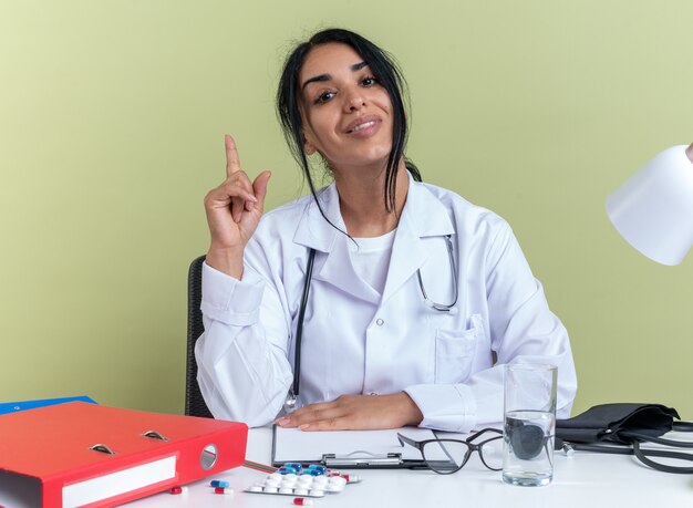 Impresionada joven doctora vistiendo bata médica con estetoscopio se sienta en el escritorio con herramientas médicas aisladas en la pared verde oliva