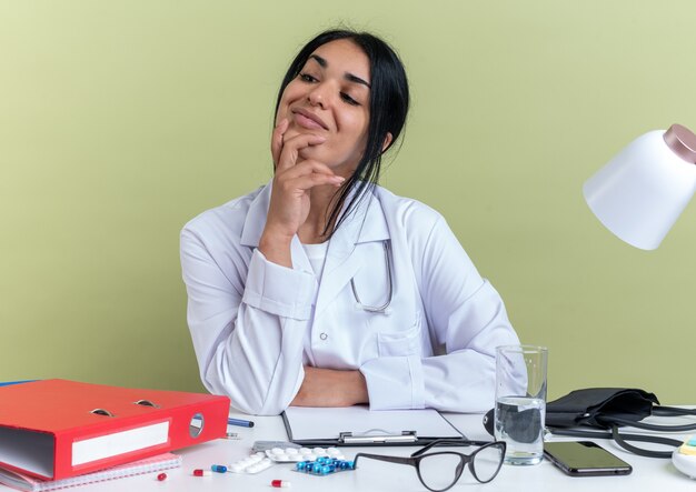 Impresionada joven doctora vistiendo bata médica con estetoscopio se sienta en el escritorio con herramientas médicas poniendo la mano en la barbilla aislada en la pared verde oliva