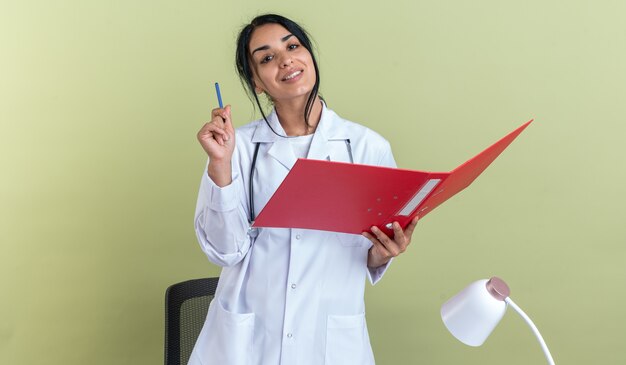 Impresionada joven doctora vistiendo bata médica con estetoscopio sosteniendo la carpeta con lápiz aislado en la pared verde oliva