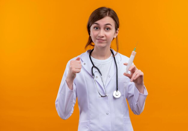 Impresionada joven doctora vistiendo bata médica y estetoscopio sosteniendo una jeringa con el puño cerrado en un espacio naranja aislado con espacio de copia