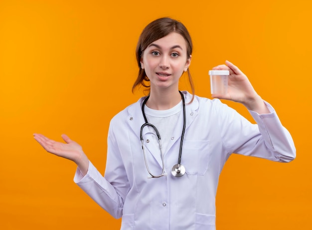 Impresionada joven doctora vistiendo bata médica y estetoscopio sosteniendo el vaso y mostrando la mano vacía en el espacio naranja aislado