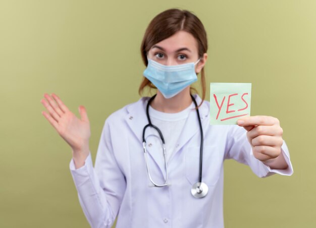 Impresionada joven doctora vistiendo bata médica, máscara y estetoscopio estirando la nota de sí y mostrando la mano vacía en el espacio verde aislado