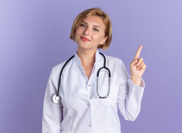 Impresionada joven doctora vistiendo bata médica con puntos de estetoscopio en aislado en la pared azul