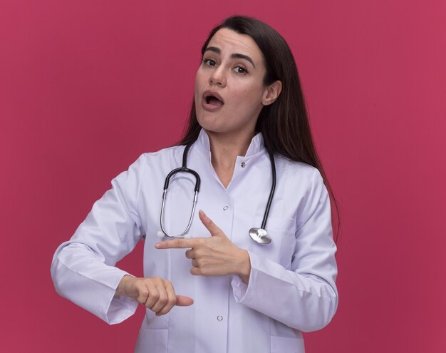 Impresionada joven doctora vistiendo bata médica con puntos de estetoscopio a los lados en rosa