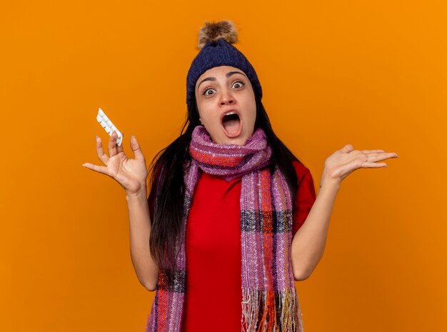 Impresionada joven enferma caucásica vistiendo gorro de invierno y bufanda sosteniendo un paquete de tabletas mostrando la mano vacía mirando a cámara aislada sobre fondo naranja