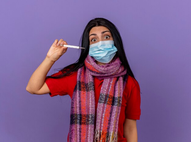 Impresionada joven enferma caucásica vistiendo máscara y bufanda sosteniendo el termómetro horizontalmente aislado en la pared púrpura con espacio de copia