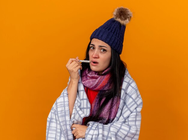 Foto gratuita impresionada joven enferma con gorro de invierno y bufanda envuelta en cuadros poniendo el termómetro en la boca mirando al frente aislado en la pared naranja