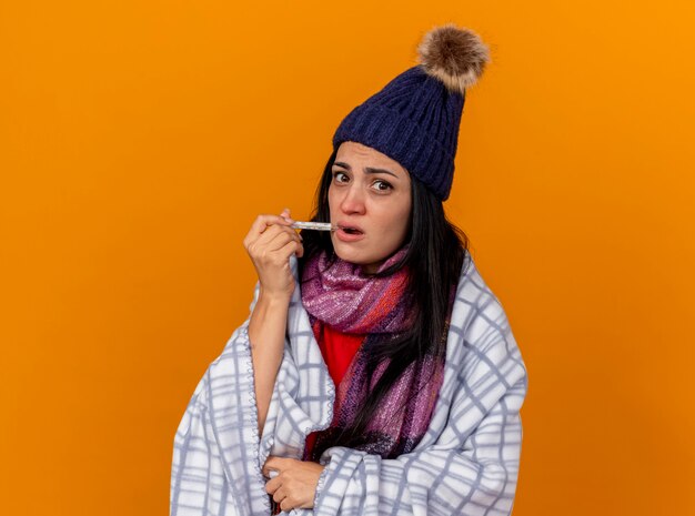 Impresionada joven enferma con gorro de invierno y bufanda envuelta en cuadros poniendo el termómetro en la boca mirando al frente aislado en la pared naranja