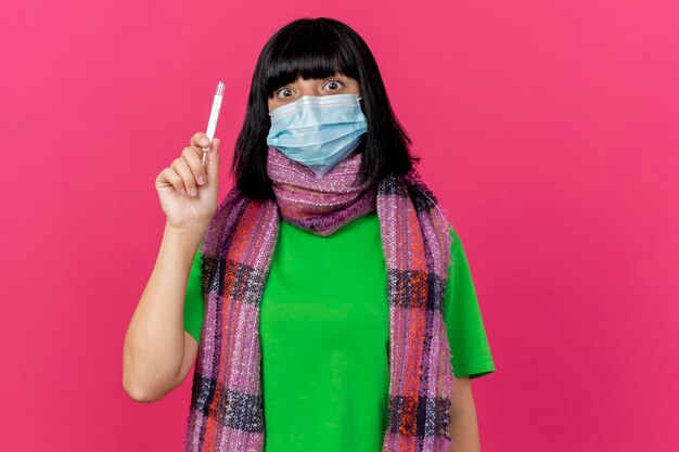 Impresionada joven enferma con máscara y bufanda sosteniendo el termómetro mirando al frente aislado en la pared rosa con espacio de copia