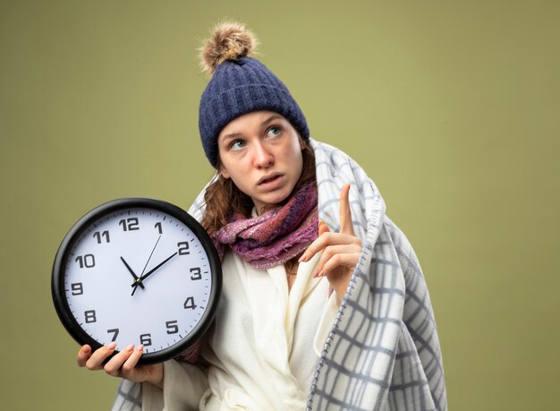 Impresionada joven enferma mirando hacia arriba vistiendo túnica blanca y gorro de invierno con bufanda sosteniendo el reloj de pared envuelto en puntos de cuadros en arriba