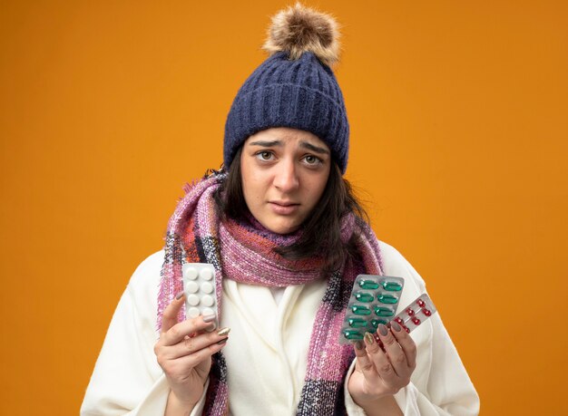 Impresionada joven enferma vistiendo bata gorro de invierno y bufanda sosteniendo paquetes de píldoras médicas mirando al frente aislado en la pared naranja