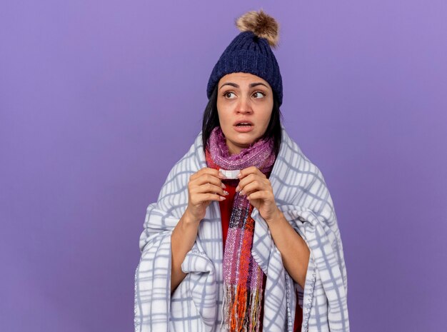 Impresionada joven enferma vistiendo gorro de invierno y bufanda envuelta en plaid sosteniendo yeso médico mirando al lado aislado en la pared púrpura