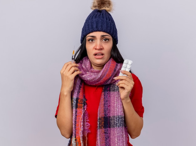 Impresionada joven enferma vistiendo gorro de invierno y bufanda sosteniendo una jeringa y un paquete de tabletas mirando al frente aislado en la pared blanca