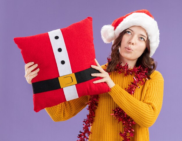 Impresionada joven eslava con gorro de Papá Noel y con guirnalda alrededor del cuello sosteniendo una almohada decorada mirando hacia arriba aislado sobre fondo púrpura con espacio de copia