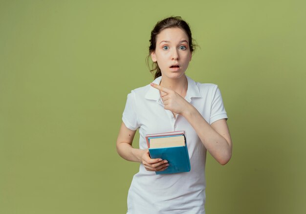 Impresionada joven estudiante bastante femenina sosteniendo el libro y el bloc de notas apuntando al lado aislado sobre fondo verde oliva con espacio de copia