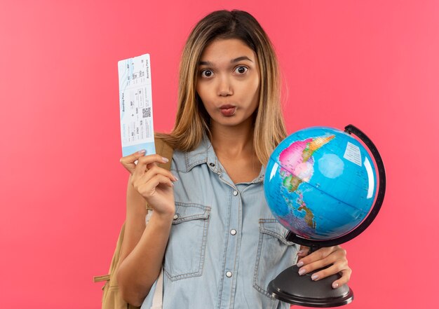 Impresionada joven estudiante bonita con bolsa trasera con boleto de avión y globo aislado en rosa