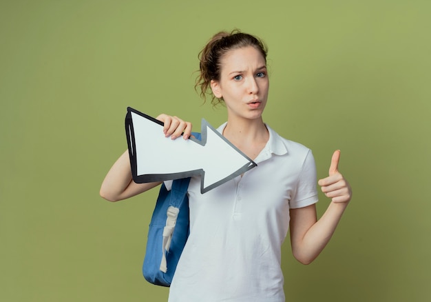 Impresionada joven estudiante bonita con bolsa trasera con marca de flecha que apunta a un lado y muestra el pulgar hacia arriba aislado sobre fondo verde oliva con espacio de copia