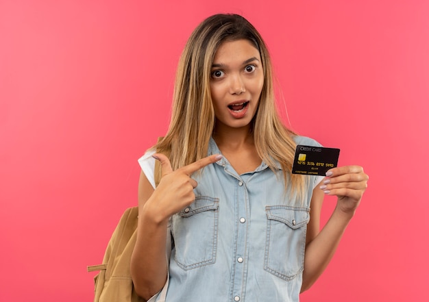 Foto gratuita impresionada joven estudiante bonita con bolsa trasera mostrando y apuntando a la tarjeta de crédito aislada en rosa