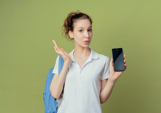 Impresionada joven estudiante bonita con bolsa trasera mostrando teléfono móvil y manteniendo la mano en el aire aislado sobre fondo verde con espacio de copia