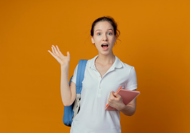 Impresionada joven estudiante bonita con bolsa trasera sosteniendo bloc de notas y bolígrafo mostrando la mano vacía aislada sobre fondo naranja con espacio de copia