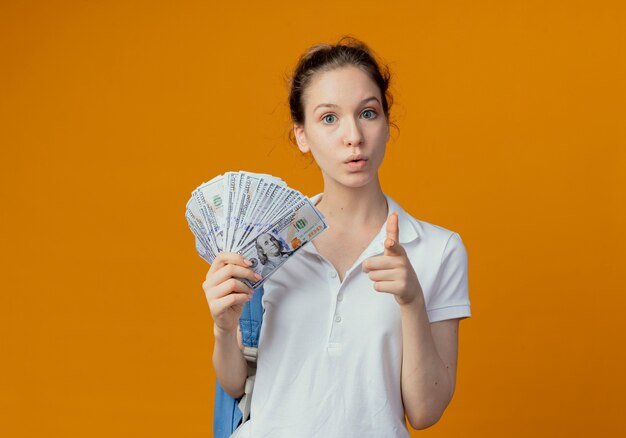 Impresionada joven estudiante bonita con bolsa trasera sosteniendo dinero y apuntando a la cámara aislada sobre fondo naranja con espacio de copia