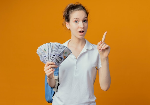 Impresionada joven estudiante bonita con bolsa trasera sosteniendo dinero y levantando el dedo aislado sobre fondo naranja con espacio de copia