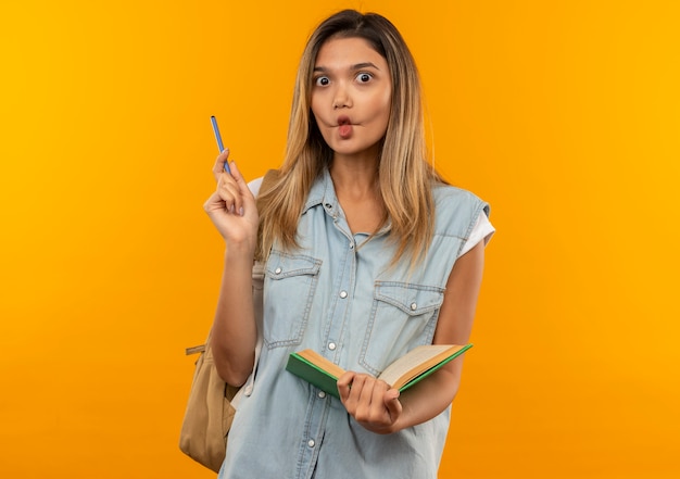 Impresionada joven estudiante bonita con bolsa trasera sosteniendo libro abierto y bolígrafo aislado en naranja