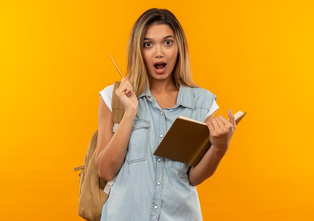 Impresionada joven estudiante bonita con bolsa trasera sosteniendo libro abierto y bolígrafo aislado en naranja