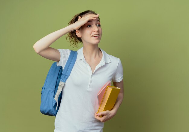 Impresionada joven estudiante bonita con bolsa trasera sosteniendo libro y bloc de notas mirando a distancia aislada sobre fondo verde oliva con espacio de copia