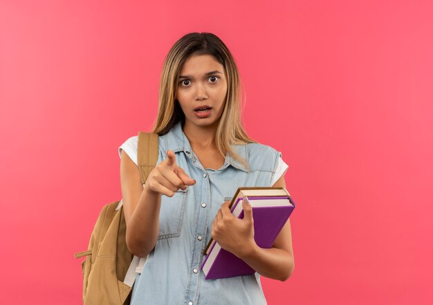 Impresionada joven estudiante bonita con bolsa trasera sosteniendo libros y apuntando al frente aislado en rosa