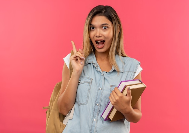 Impresionada joven estudiante bonita con bolsa trasera sosteniendo libros y levantando el dedo aislado en rosa