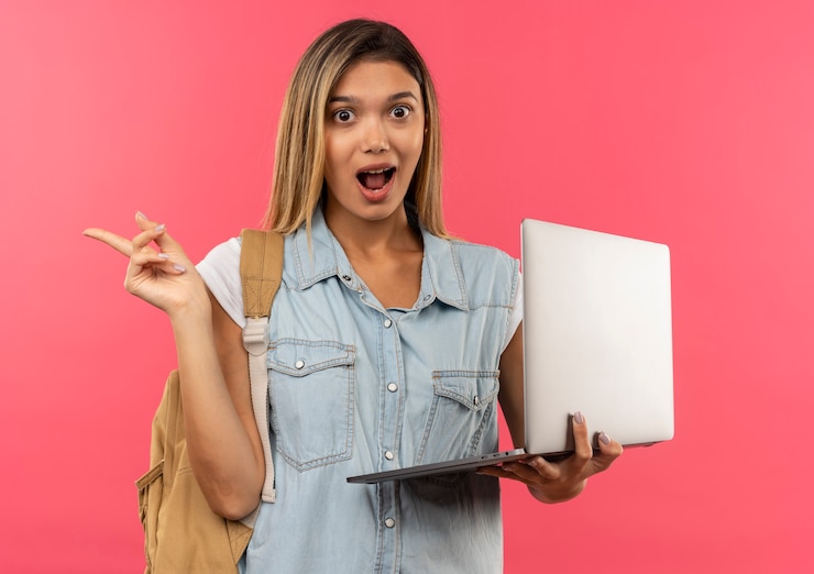 Foto gratuito impresionada joven estudiante bonita con bolsa trasera sosteniendo portátil y apuntando al lado aislado en rosa