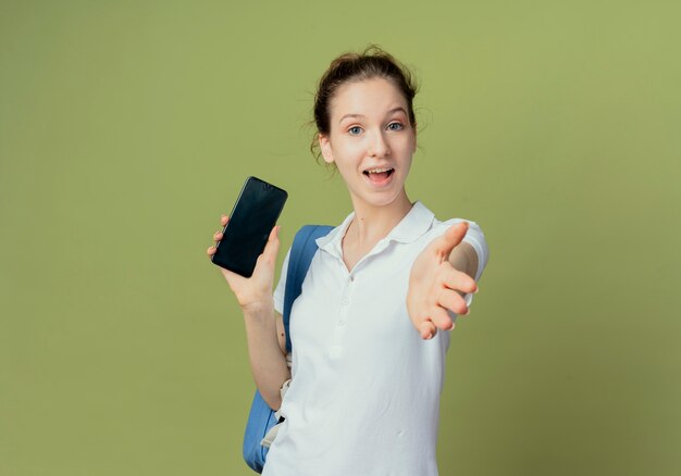 Impresionada joven estudiante bonita con bolsa trasera sosteniendo el teléfono móvil y estirando la mano