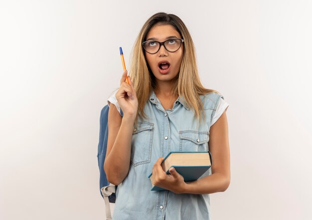 Impresionada joven estudiante bonita con gafas y bolsa trasera con libro y bolígrafo mirando hacia arriba aislado en blanco