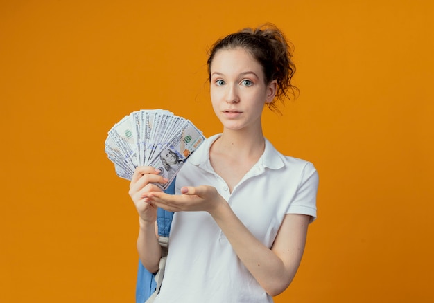 Impresionada joven estudiante bonita con mochila sosteniendo y apuntando con la mano al dinero aislado sobre fondo naranja con espacio de copia